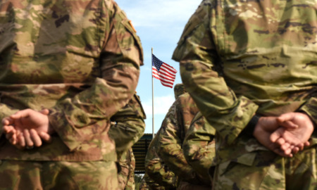 National Guard school Violence: Closeup of backs of people standing in two lines wearing camo green and brown army uniforms with an American flag in background