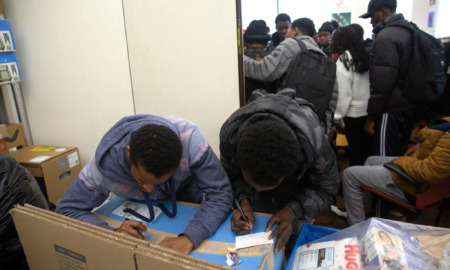 NYC homeless youth and migrants: Two black males fill out forms using the tops of large cardboard boxes for desks while several other people stand crowded together in another room in the background