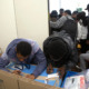 NYC homeless youth and migrants: Two black males fill out forms using the tops of large cardboard boxes for desks while several other people stand crowded together in another room in the background