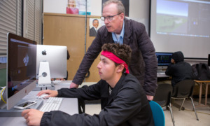 Tutoring Improves Attendance: Older gray-haired man with glasses in dark suit stands beside seated teen male student with red headband and dark jacket, both looking at large monitor in computer lab