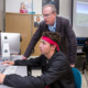 Tutoring Improves Attendance: Older gray-haired man with glasses in dark suit stands beside seated teen male student with red headband and dark jacket, both looking at large monitor in computer lab