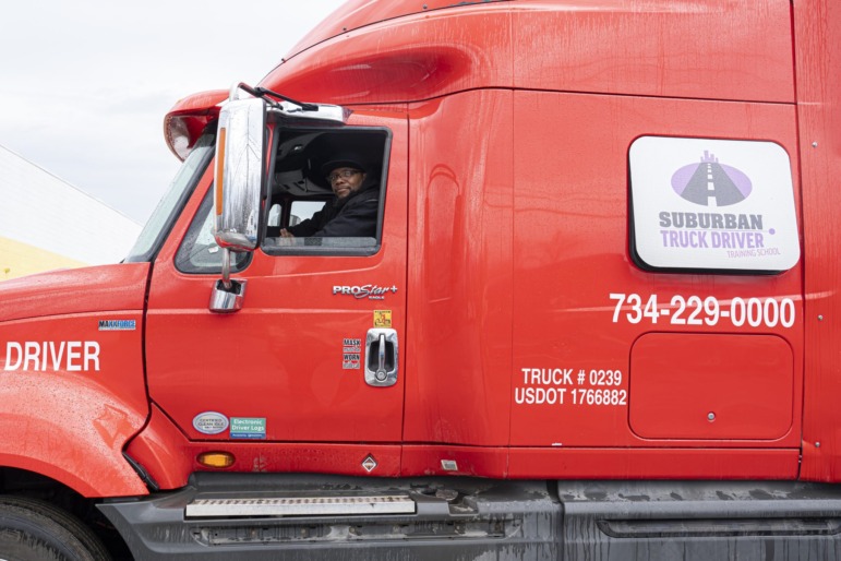 Prison No Education: Black man in dark winter jacket, hat sits inside cab at driver's wheel of red semi-truck.