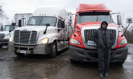 Prison No Education: Black man in dark winter jacket, hat and pants stands leaning against front of bright red semi-truck parked next to a white semi-truck on asphalt parking lot under gray, rainy sky