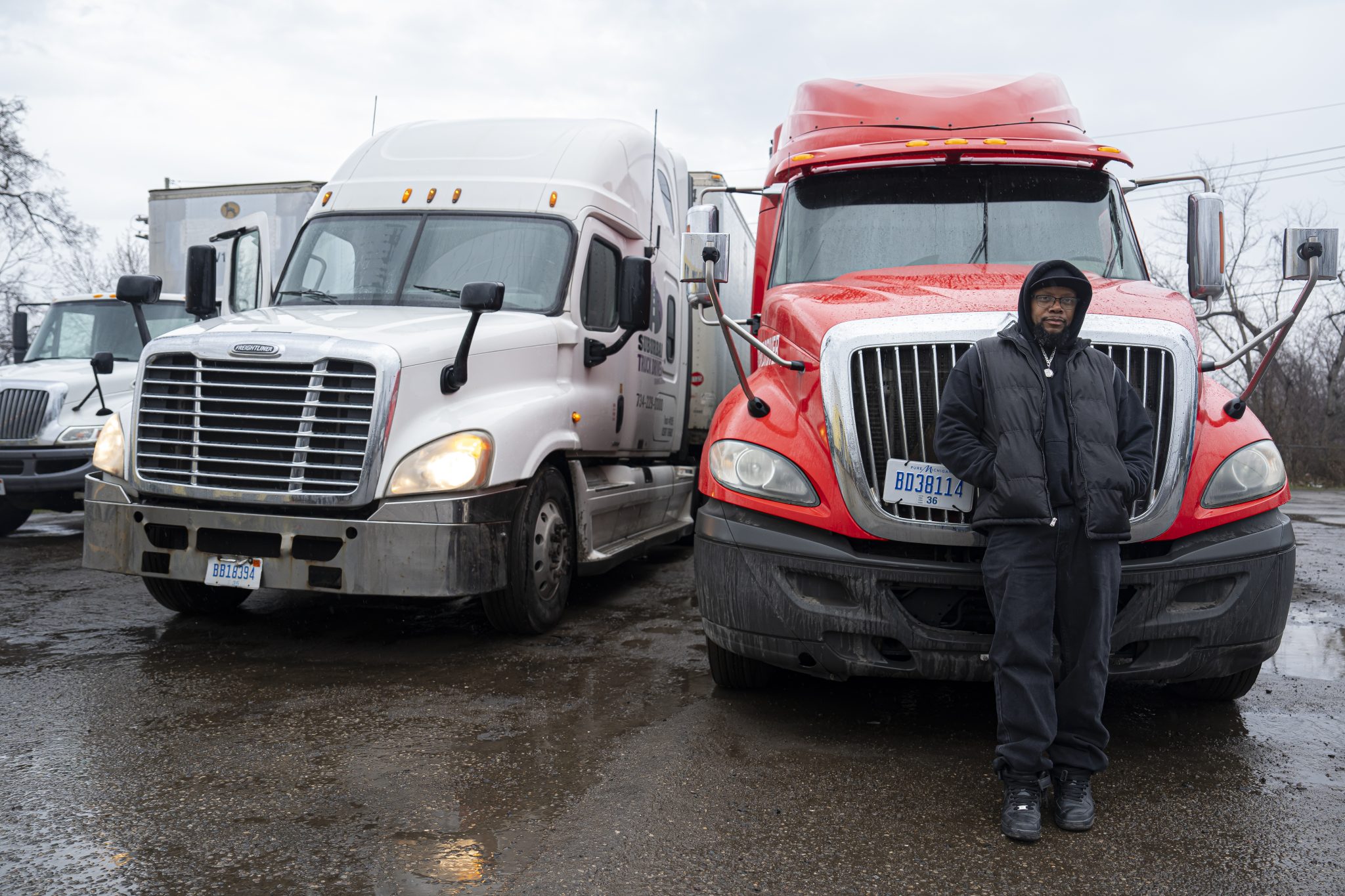 Prison No Education: Black man in dark winter jacket, hat and pants stands leaning against front of bright red semi-truck parked next to a white semi-truck on asphalt parking lot under gray, rainy sky