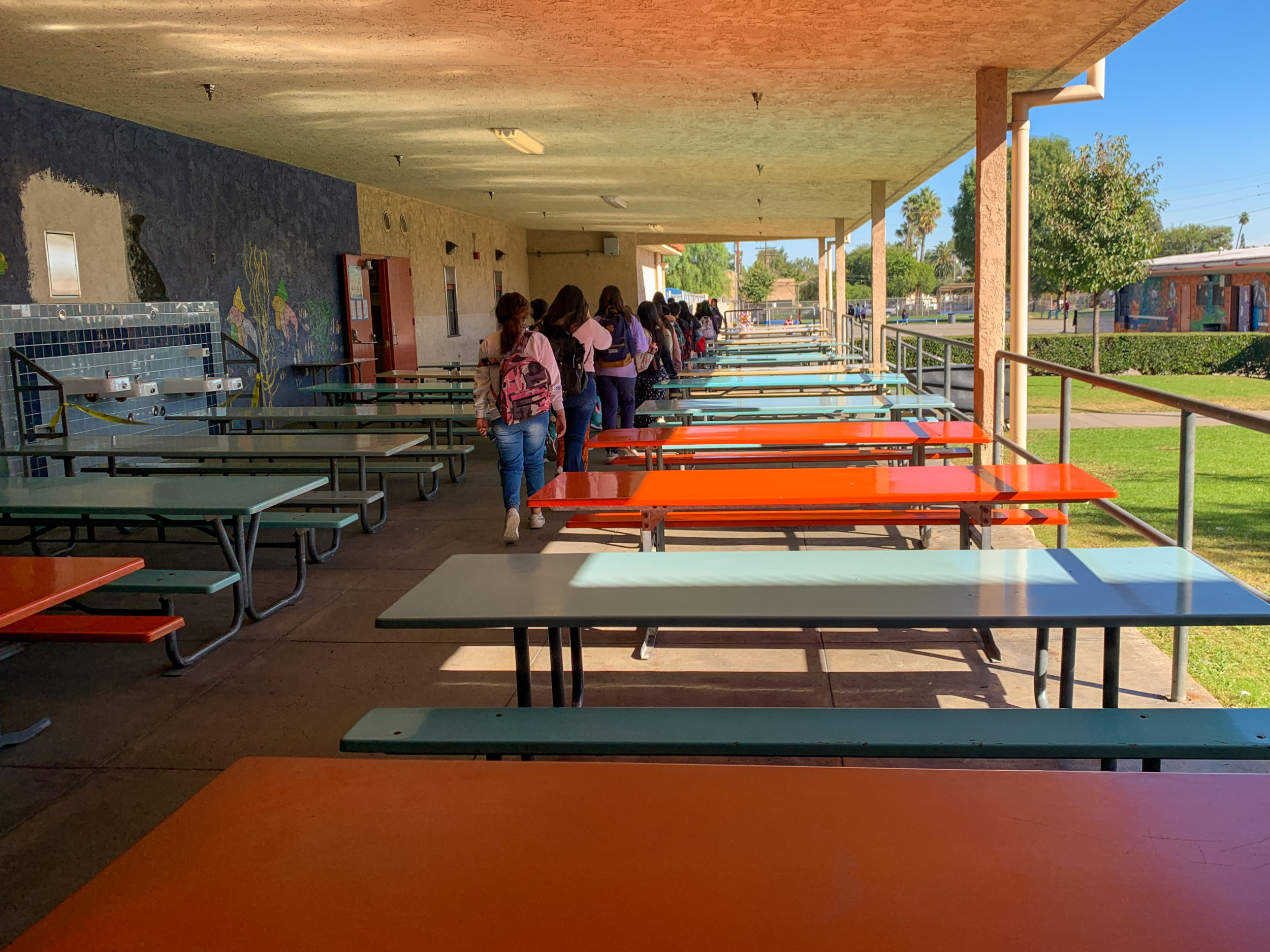 No school suspension: Two rows of many empty, long tables on pale blue or bright orange with attached benches are in a covered cemented area, with several people lined up single file in aisle between them