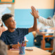 Tutoring analysis: Young Black male teen in navy t-shirt sitting at school desk gives teacher in white suit a high five