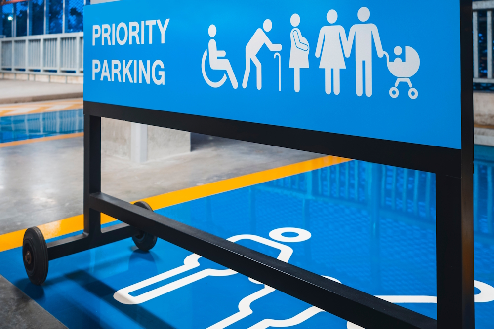 Universal Design: Bright blue metal sign on wheels in indoor parking lot, with white text "Priority Parking" and 6 white disability icons