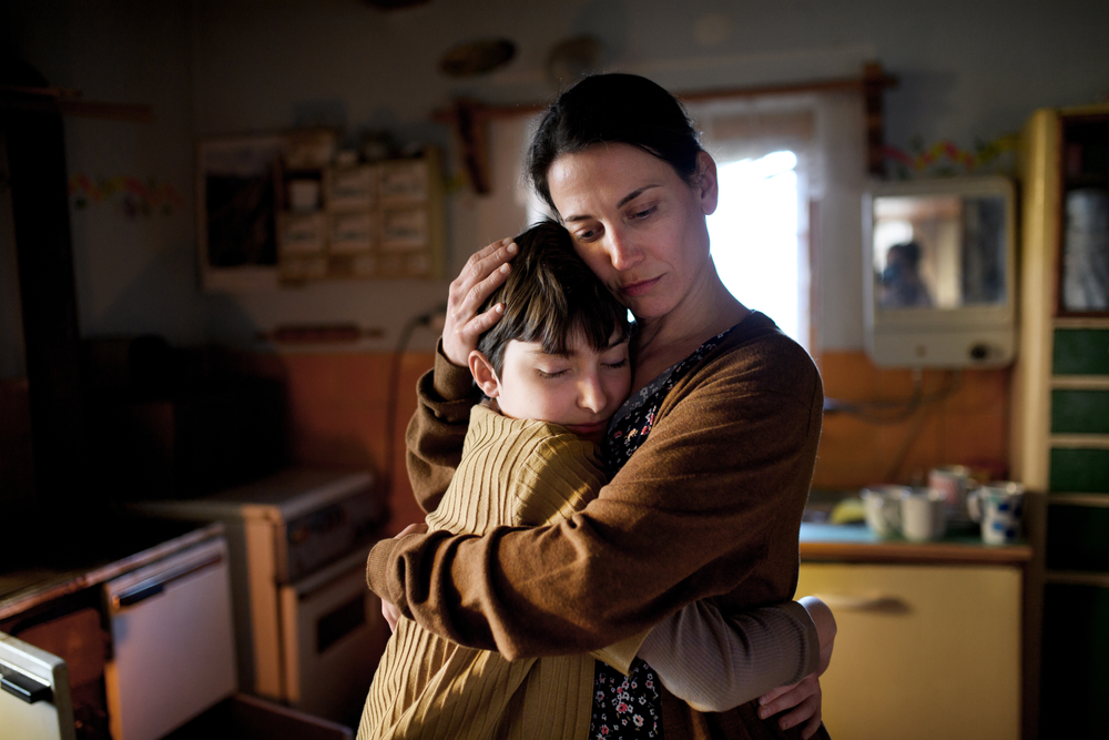 Child support payments: Portrait of sad poor mature mother with dark hair pulled back in brown sweater, standing hugging preteen daughter indoors at home in older, dark kitchen.