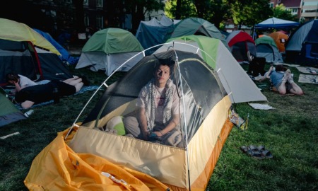 College protest photo essay: Young man with dark hair sits inside small yellow and white dome tent on a green grass lawn surrounded by similar tents and young adults