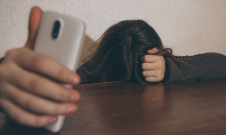 Survey teen depression and smart phones: Person with long dark hair sits with head down on dark brown table top hiding face and holding silver smart phone in right hand with arm on table reaching towards camera