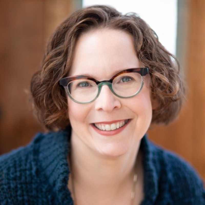 Teen survey depression: Headshot smiling woman with short brown hair and dark framed glasses in navy blue top.