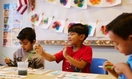 Colorado public preschool: Three preschool bots all with dark fair sit at a long table in front of a colorful bulletin board water coloring