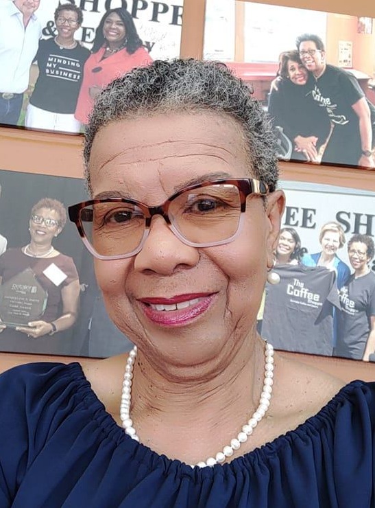 Segregation-Academies: Headshot Black woman with short, salt and pepper hair and dark glasses wearing navy top sitting in front of a corkboard with several photos