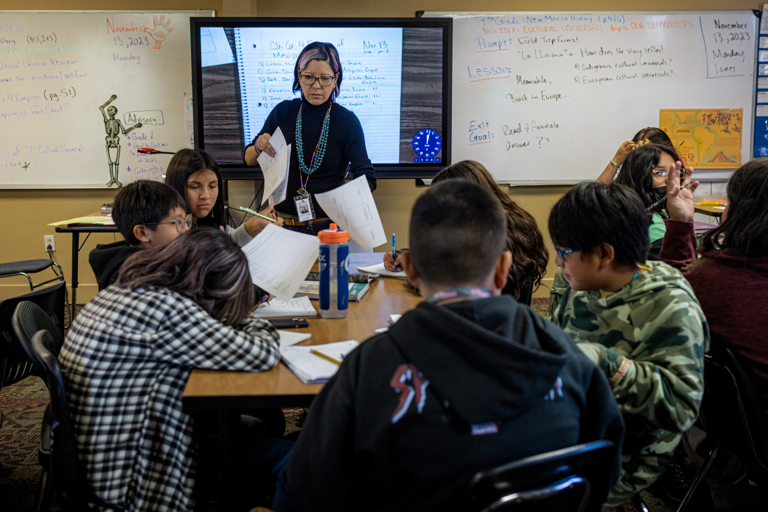 Native American Charter School; Woman teacher stands facing several students seated around a long table