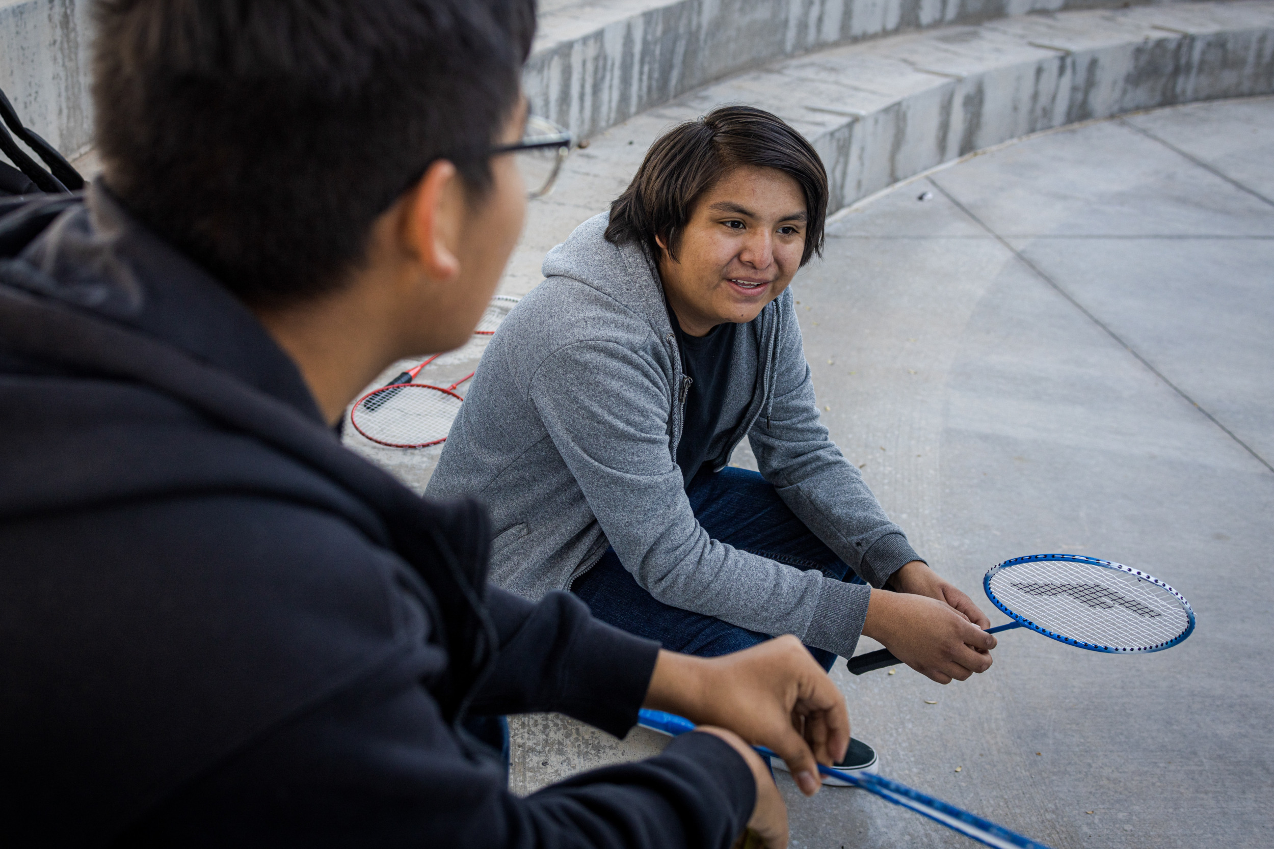 Native American Charter School; Two dark-haired male teens sit outside on cement steps in long-sleeved shorts and jeans