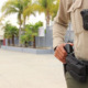 Police on campus: closeup of only middle and right side body showing hand on gun in holster of police on tan and brown uniform with modern high school campus in the background