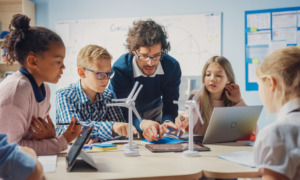 School skills research: Male teacher leans over table with 5 elementary students explaining a science windmill model.