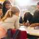 Teacher layoffs: Group of elementary school kids stand around a table in a classroom, attentively leaning over table and following a lesson being taught by their male teacher with glasses, dark hair and beard.