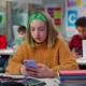 School cell phone bans: Teenage schoolgirl with green hair chatting on smartphone sitting at desk in class. Teen student using cellphone in classroom at school