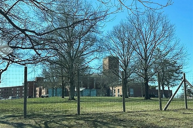 Shrub Oak School investigation: Large buildings across a wide green lawn seen through trees and a tall wire fence