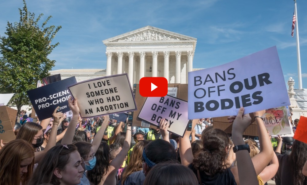 Abortion bans: Crowd of adults at rally with handmaid signs held up gathered in front of multi-story, white building with 8 pillars in front