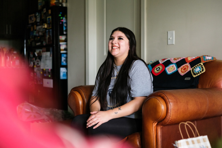 Older Immigrant student school admissions: Smiling Latinx girl with very long black hair in gray t-shirt sits in over-stuffed brown leather chair in her living room looking up and smiling