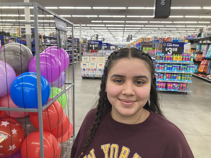 Older Immigrant student school admissions: Headshot of young Latino woman with long dark hair in a maroon sweatshirt standing inside a Walmart with aisles of merchandise behind her and a tall wire display basket fill of colorful balls next to her