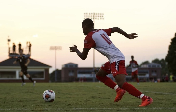 Older Immigrant student school admissions: Black male athlete in red and white uniform suns on grass field after a black and white soccer ball