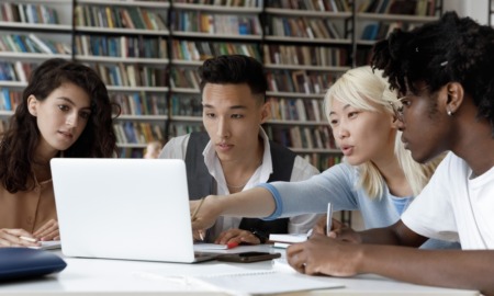 College Essay after affirmative action ended: Four multiethnic schoolmates look at laptop in library discussing information they see on screen