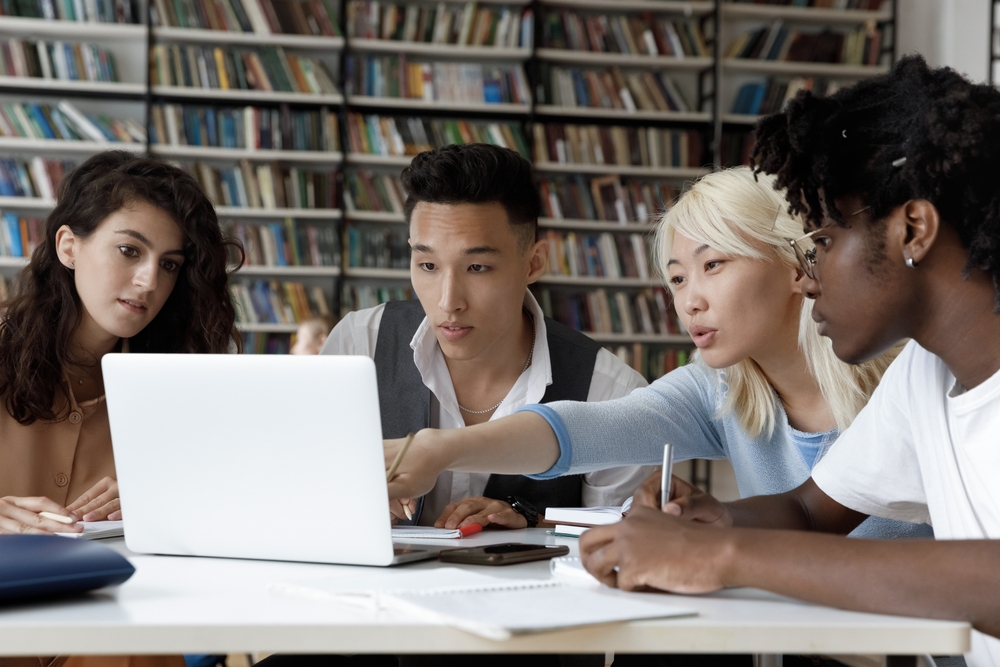 College Essay after affirmative action ended: Four multiethnic schoolmates look at laptop in library discussing information they see on screen