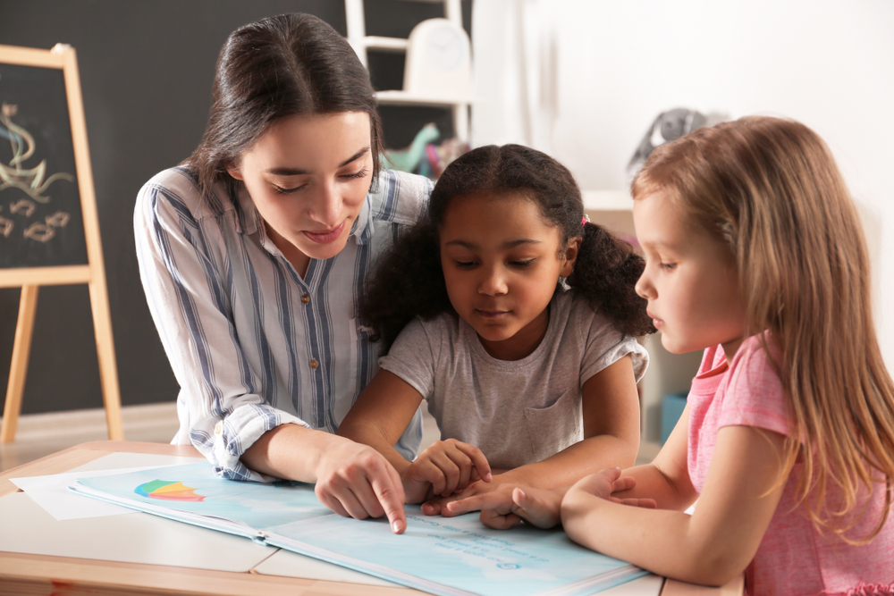 K-3-literacy-scores: Two young girls sit at desk with teacher reading a book.