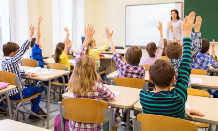 Segregation academies: Back view of group of all white school kids sitting at classroom desks and raising hands with white female teacher standing facing students