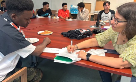 Older Immigrant students high school enrollment: Group of Black, male, older teens sit stound wood conference table with older brown-haired white woman helping one in foreground to fill out paperwork