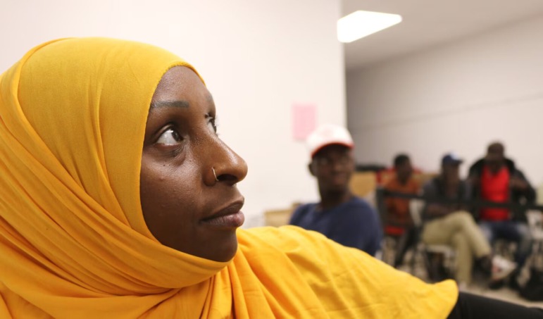 Older Immigrant students high school enrollment: Closeup of profile of niddle-aged Black woman n bright yellow hajib with four Black males seated in chairs in background