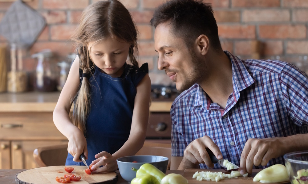 Math talk by parents to children: Adult man with short brown hair in blue plaid shirt talks to young girl with long blonde hair in navy dress as both stand at kitchen counter chopping vegetables