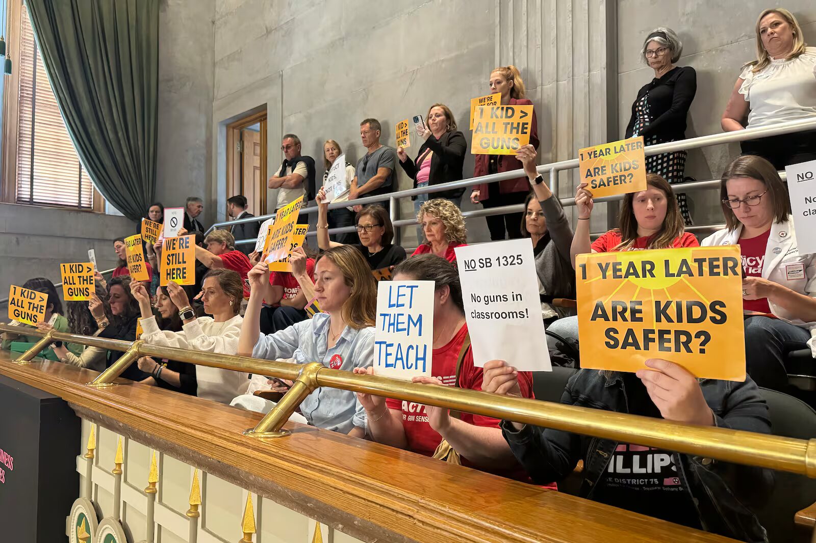 Tennessee arms teachers: Several adults stand and sit in balcony gallery area, many holding signs with language protesting arming teachers in schools