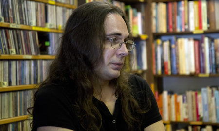 trans: Person with long brown hair, glasses, short-sleeved black top looks thoughtful sitting in bookstore