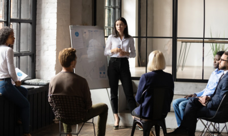 Nonprofit funding goals: Young woman with long dark hait stands at whiteboard talking to several people seated in half -circle in front of her