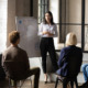 Nonprofit funding goals: Young woman with long dark hait stands at whiteboard talking to several people seated in half -circle in front of her