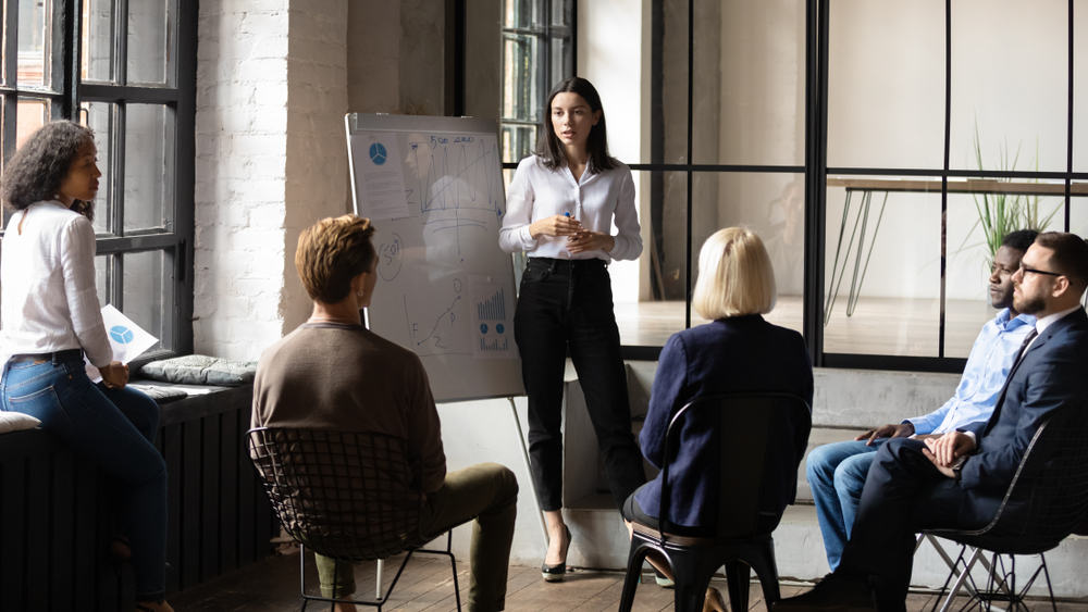 Nonprofit funding goals: Young woman with long dark hait stands at whiteboard talking to several people seated in half -circle in front of her