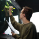 AI & education: Man with brown hair and black glasses in brown long-sleeved top stands with back to camera writing with yellow highlighter on glass blackboard, holding several papers in other hand.