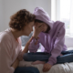 Child Anxiety Treatment: Asult woman with short brown hair crouches next to young teen chile sitting cross-legged in bed covering face with hand wearing lavendar hoodie with hood up and blue jeans