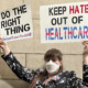 Transgender laws: Person in facemask with brown hair and glasses wearing black and white print jacket holds up two large signs "Do the right thing" and "Keep hate out of healthcare"