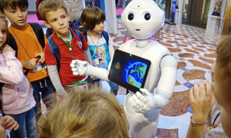 Robots in schools: White, child-sized, humanoid robot with computer monitor embedded in chest showing picture of earth, stands with hands outreached to group of elementary students surrounding it