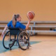 Adaptive sports: Teen with long blonde ponytail sitting in wheelchair on indoor basketball court holds basketball up in air in right hand