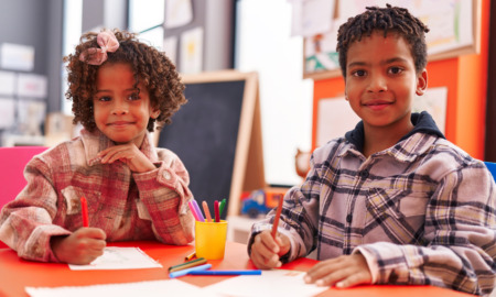 Black charter school: Two young Black children in plaid shirts sit next to each other at a school desk drawing on paper smiling into camera