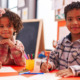 Black charter school: Two young Black children in plaid shirts sit next to each other at a school desk drawing on paper smiling into camera