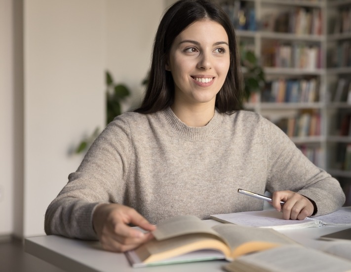 Literacy and touch: Teen girl with long dark hair in off-white sweater sits in library writing a paper while reading books.