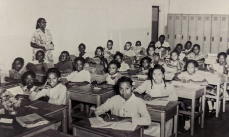 Classroom of all Black students seated in 4 rows of desks with female Black woman teacher standing against wall to left of desks