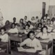 Classroom of all Black students seated in 4 rows of desks with female Black woman teacher standing against wall to left of desks